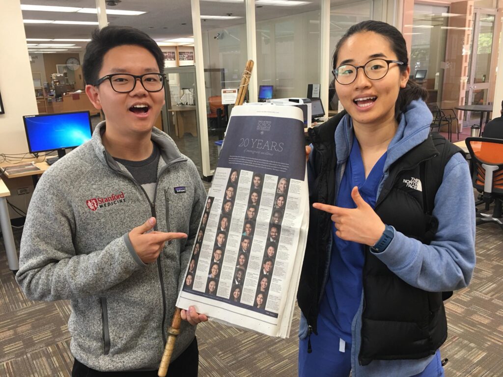 Photograph of two people in their 20s holding a New York Times and pointing at their own pictures. 