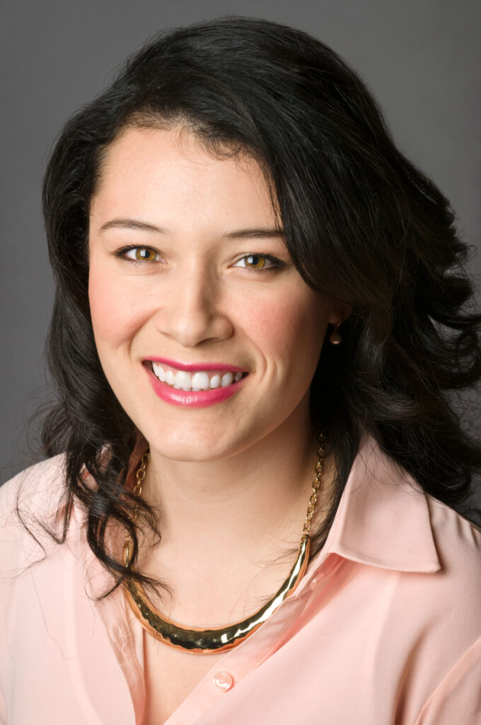 Headshot of a woman in her 20s who has heritage from Mexico with light skin tone and black curled hair with a side part. She is wearing a light pink button up, gold chunky necklace and smiling at the camera.