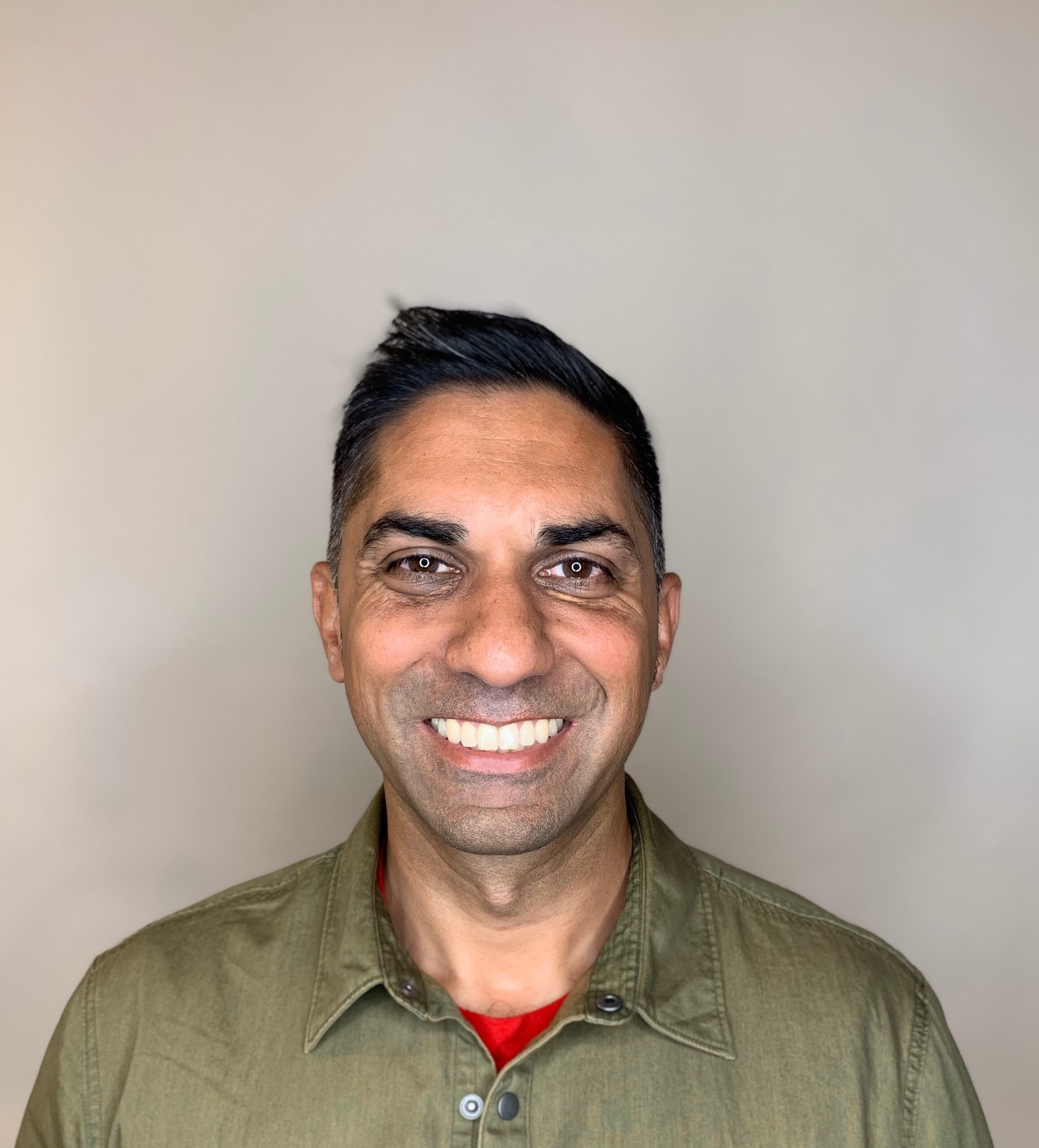 Headshot of a man in his 40s who has heritage from India with light-medium skin tone and black short hair. He is wearing an army green button up and smiling at the camera.