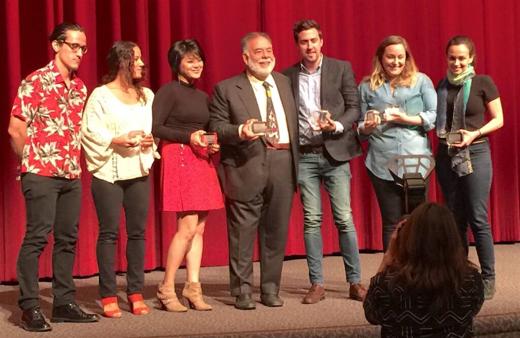 Photograph of seven people standing in front of a red curtain on a stage holding small awards. 