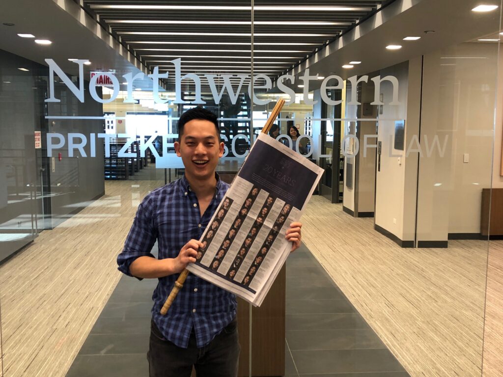 Photograph of a man in his 20s who has heritage from China and Taiwan with light skin tone and short black hair. He is holding a New York Times in front of a glass door that says "Northwestern Pritzker School of Law".