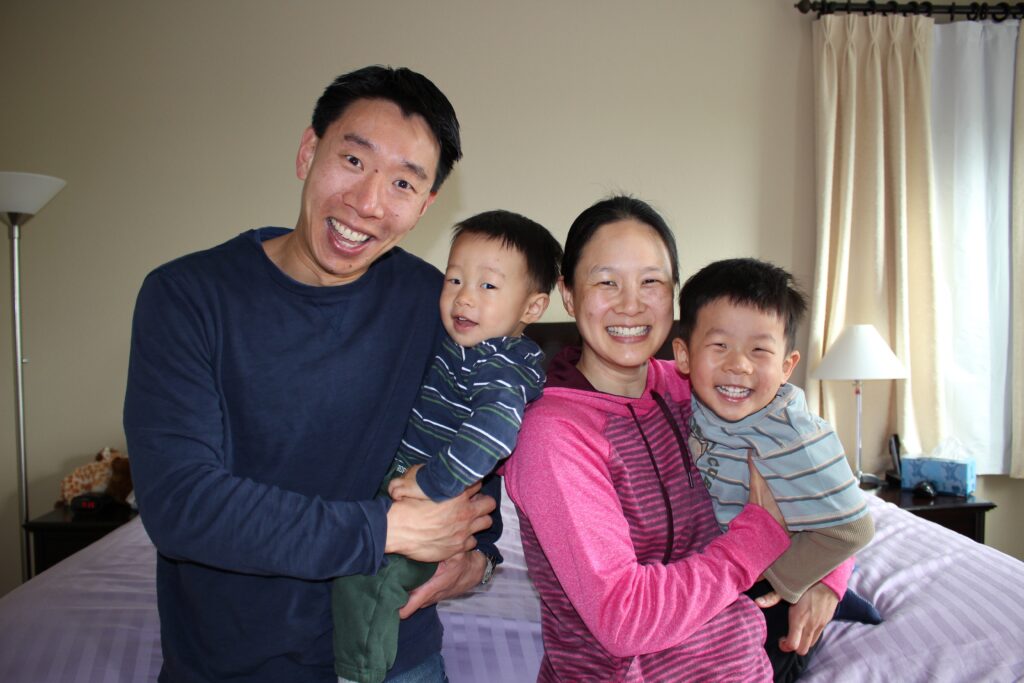 Photograph of a family, dad, mom and two young children. They are standing in a bedroom and smiling at the camera. 