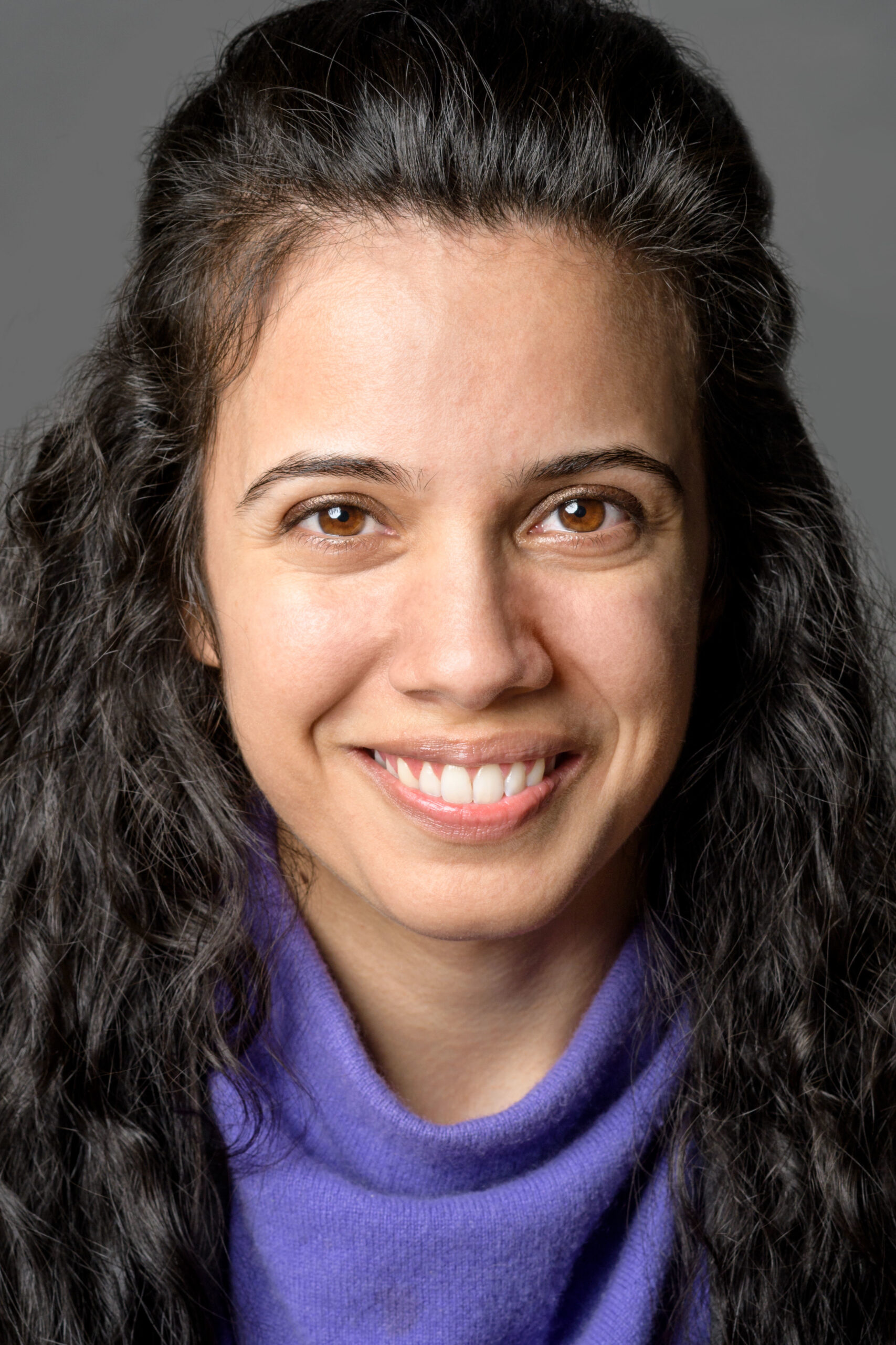 Headshot of a woman in her 20s who has heritage from Iran, she has light skin tone and curly long black hair, half pulled back. She is wearing a purple sweater and smiling at the camera.