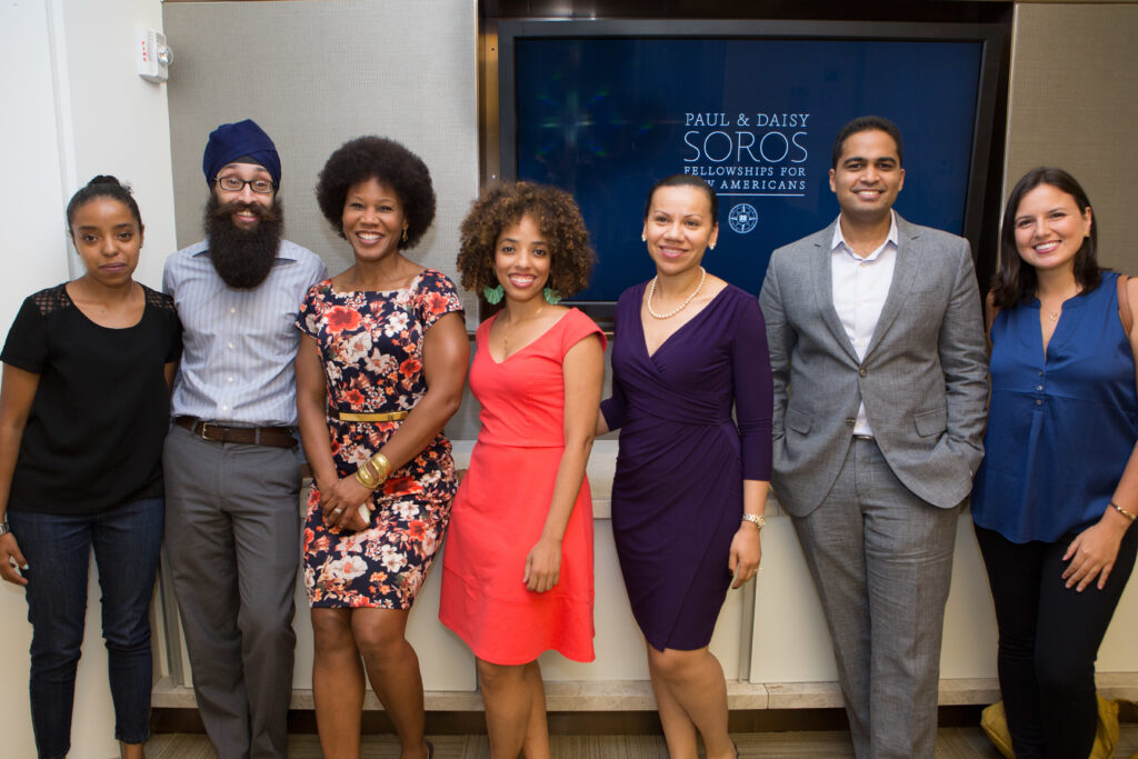 Group photograph of seven people posing together against a wall with a monitor displaying the PDSoros logo. 