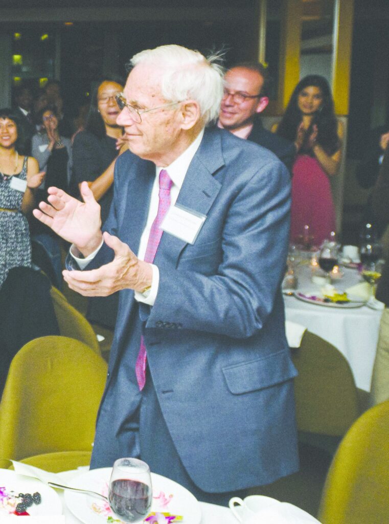 Photograph of a man in his 80s with light skin tone and white hair. He is wearing a navy suit, white button up shirt and red tie. He is looking off camera left, smiling and clapping. People can be seen behind and around him also clapping. 