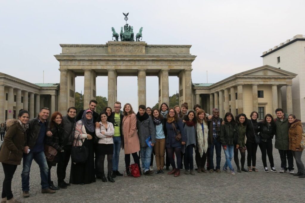 Photograph of a large diverse group of people, dressed for cooler weather, they are standing in front of a columned structure. 