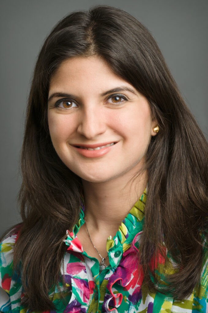 Headshot of a woman in her 20s who her heritage from Iran, with light skin tone and long dark brown hair parted. She is wearing a brightly multi-colored button up with ruffles, a gold necklace and gold stud earrings. She is smiling at the camera. 