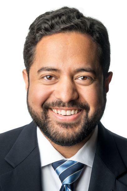 Headshot of a man in his 20s who has heritage from India with light-medium skin tone, short black wavy hair and a short-trimmed beard. He is wearing a navy suit, white button up shirt and blue tie. He is smiling at the camera. 