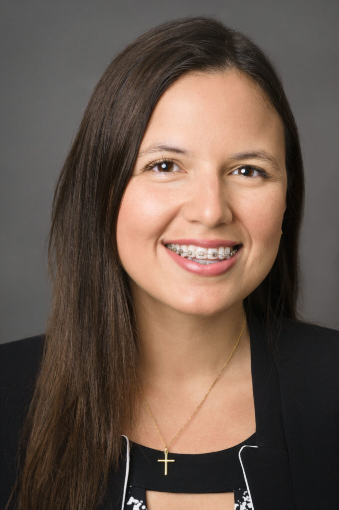 Headshot of a woman in her 20s who has heritage from Peru with light skin tone and long straight dark brown hair. She is wearing a black blazer, white and black top and a gold cross necklace. She is smiling with braces at the camera.