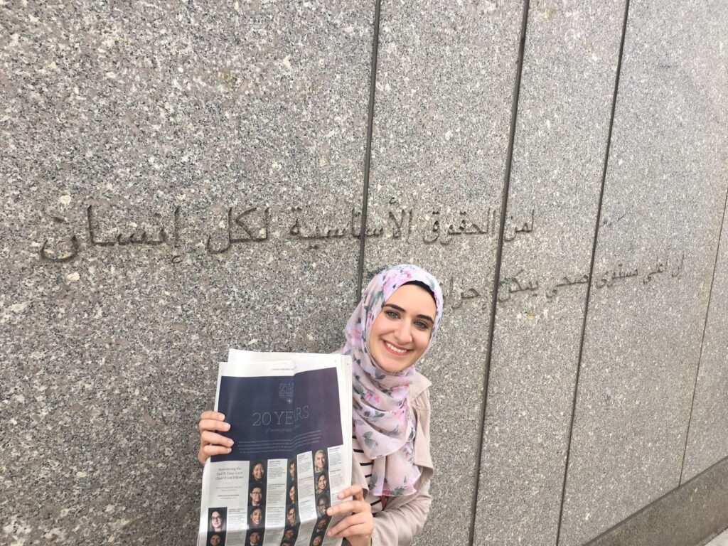 Photograph of a woman in her 20s who has heritage from Palestine with light skin tone. She is wearing a light pink floral hijab and holding a New York Times.