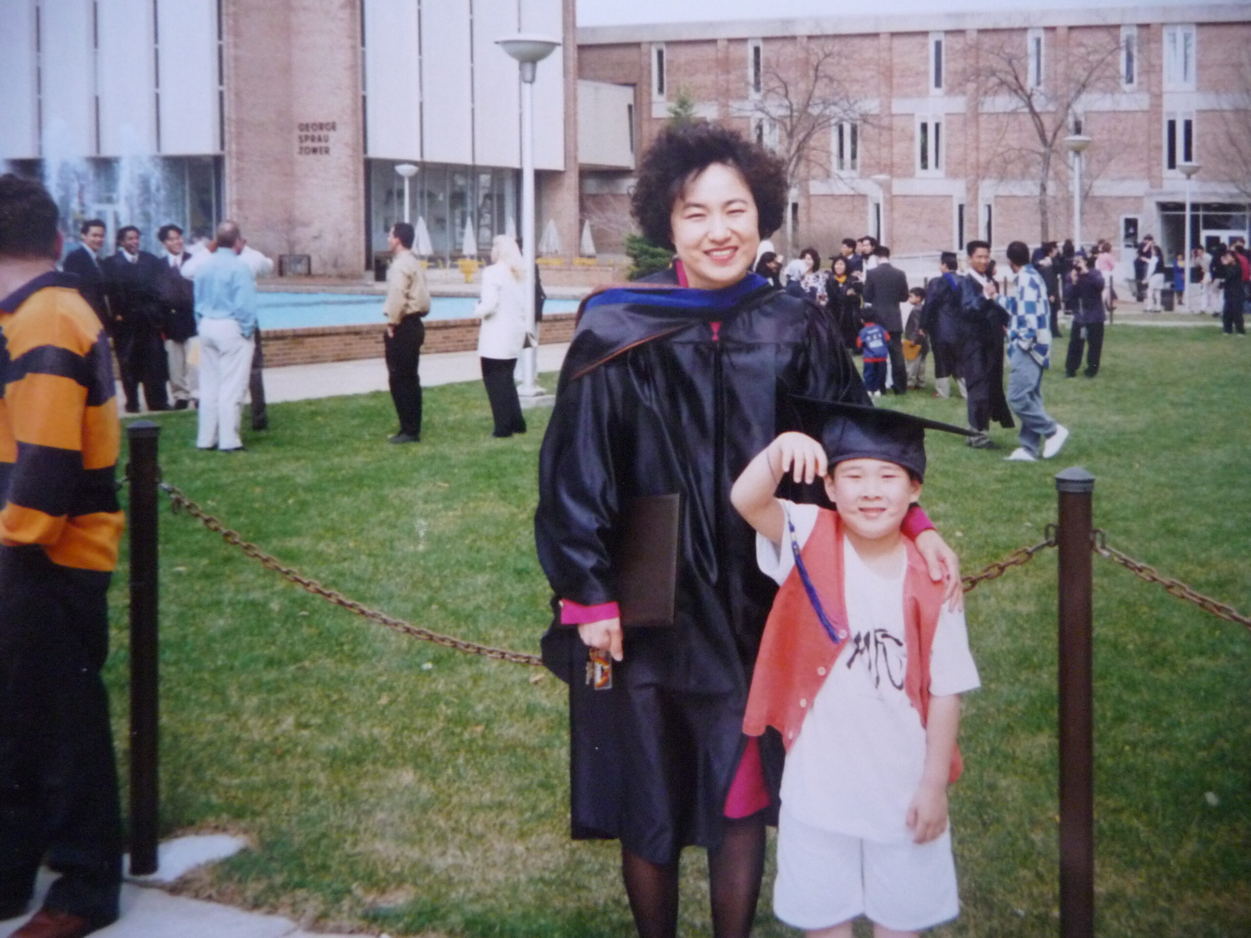 Old photograph of a woman in her 30s with light skin tone and short black hair, wearing a graduation gown, she is posed with a little boy wearing a graduation cap, white t-shirt and red vest. 