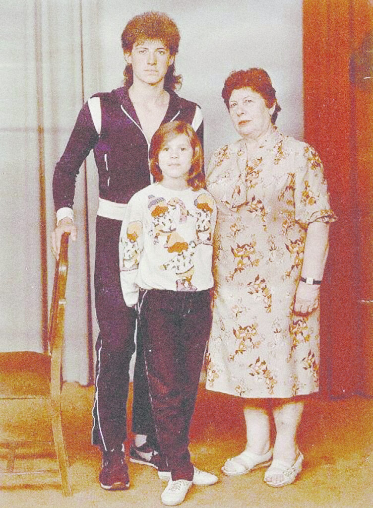 Photograph from the 1980s, three people stand looking at the camera. Foreground is a young girl about 10 wearing a sweatshirt with a graphic and has shoulder length hair, behind her a man in his teens with a mullet wearing a matching athletic set, on the right a woman in her 60s wearing a floral dress.