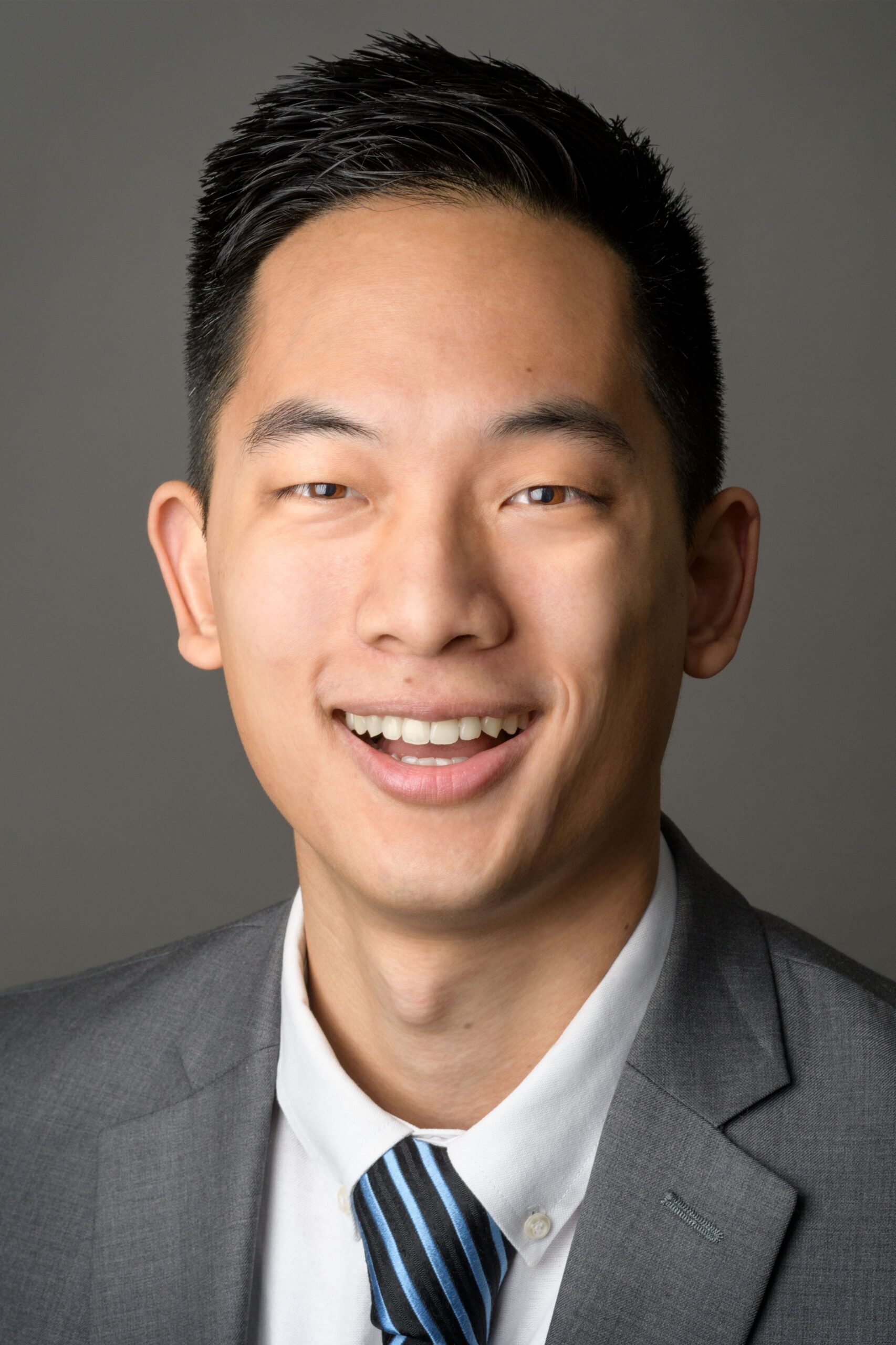 Headshot of a man in his 20s who has heritage from Taiwan with light-medium skin tone and black hair. He is wearing a grey suit, white button up and blue tie. He is smiling at the camera. 