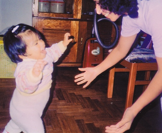 Old photograph of a woman bending down, arms open to a toddler who has her arms up and tongue out. 