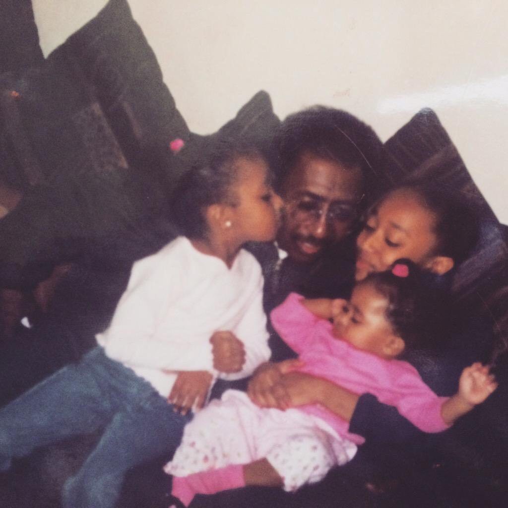 Old photograph of a man in his 30s with dark skin tone, black hair and mustache. Three young girls sit on his lap, one is giving him a kiss on the cheek. 