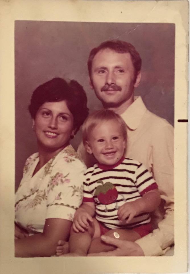 Family photograph circa 1970s, mom is wearing a white floral top, dad is wearing a button up shirt and baby is wearing a white with black stripe top with a tomato on the front. 