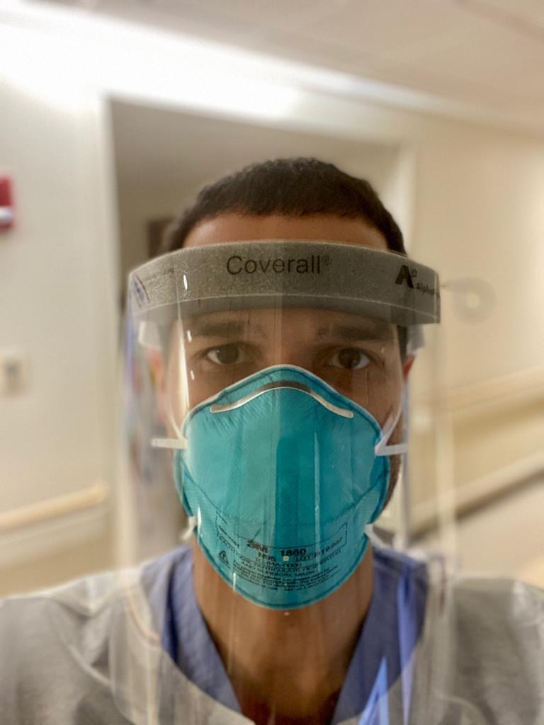 Selfie of a man in his 30s who has heritage from the Dominican Republic with light-medium skin tone and dark brown buzzed hair. He is wearing blue scrubs under full PPE (face shield, face mask, overall). He is standing in the hallway of a hospital.