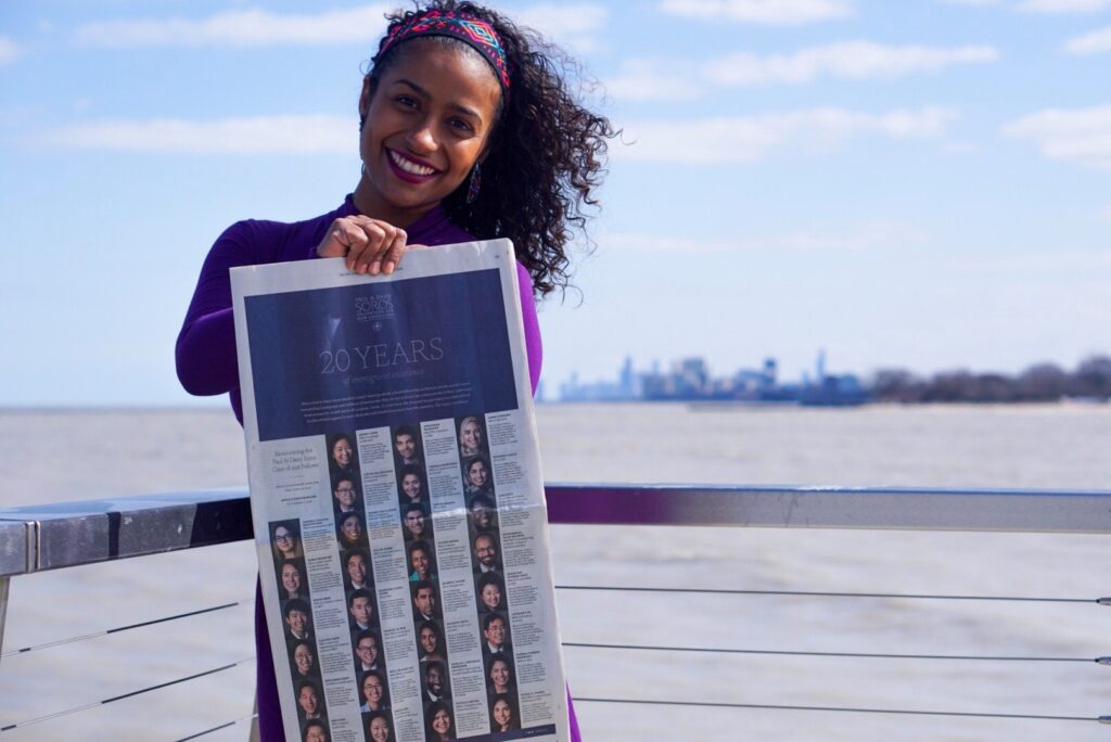 Photograph of a woman in her 20s who has heritage from Mexico with medium skin tone and curly black shoulder length hair. She has a pink and blue patterned headband on. She is holding a New York Times, a blurry skyline can be seen behind her. 