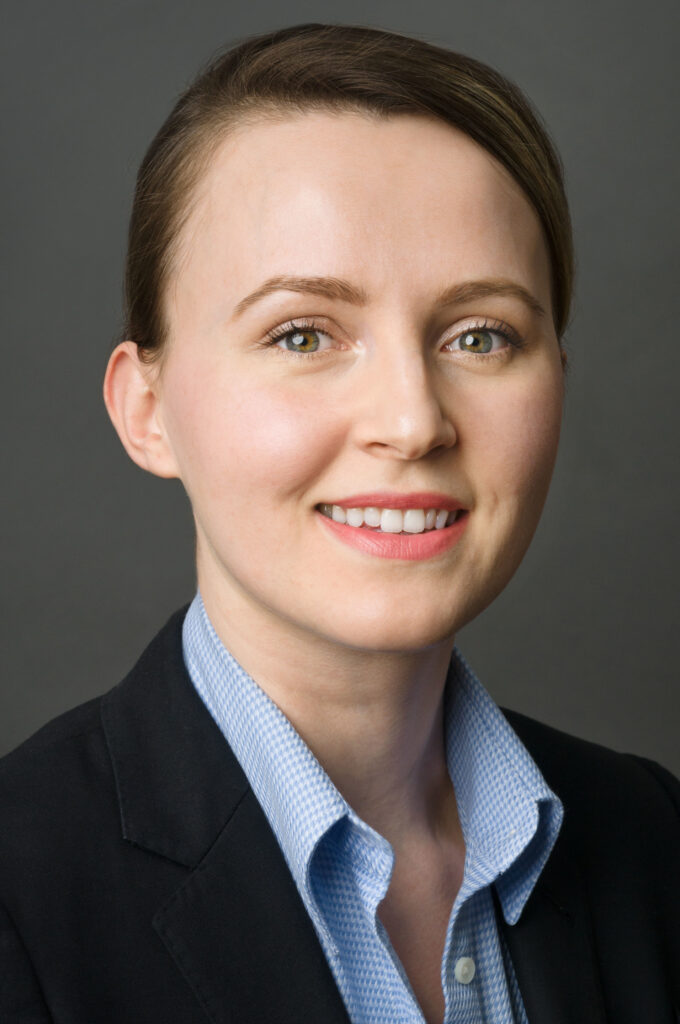 Headshot of a woman in her 20s who has heritage from Albania, with light skin tone and light brown hair parted on her right side and pulled back. She is wearing a black blazer with blue and white gingham button up shirt. She is smiling at the camera. 