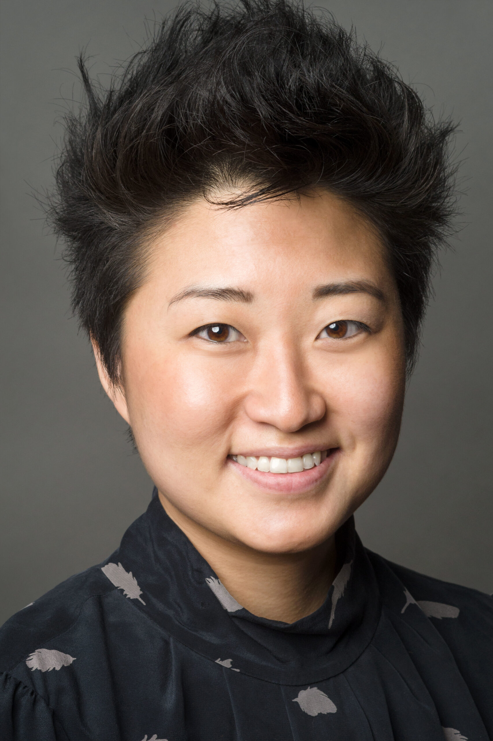 Headshot of a woman in her 20s who has heritage from South Korea with light skin tone and black pixie hair. She is wearing a black blouse, with grey patterns. She is smiling at the camera.