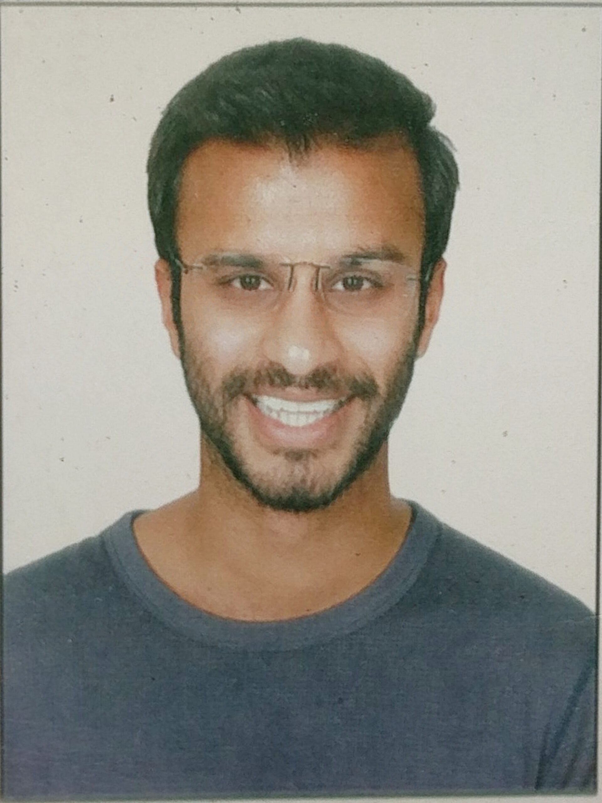 Headshot of a man in his 30s with heritage from India with medium skin tone, short black hair and a short beard. He is wearing a blue t-shirt, frameless glasses and he is smiling at the camera.