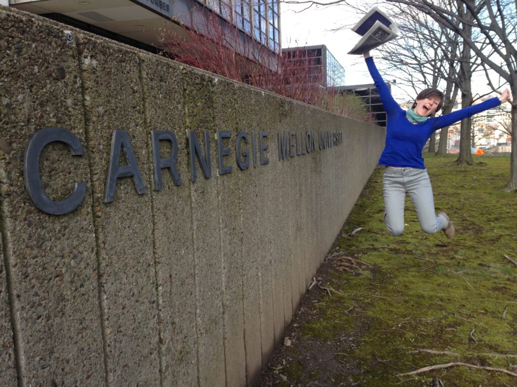 Photograph of a woman in her 20s who has heritage from Poland with light skin tone and light brown hair. She is holding a New York Times and a sign that says "Carnegie Mellon University" can be seen next to her. 