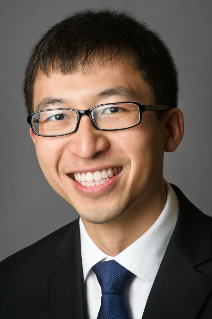 Headshot of a man in his 20s who has heritage from Myanmar with light skin tone and black Cesar cut hair. He is wearing a black suit, white button up shirt, blue tie and black rimmed narrow-rectangle glasses. He is smiling at the camera.