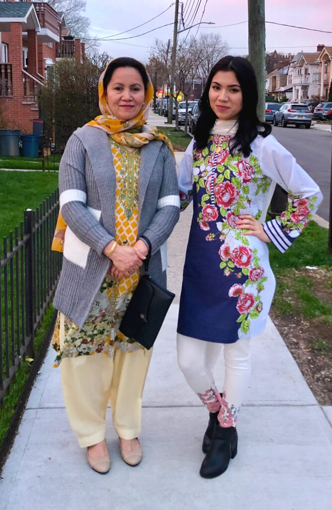 Photograph of two women standing on a sidewalk. The woman on the right is in her 20s with light skin tone and black straight long hair. She is wearing white top and pants with elaborate floral pattern on both. The woman to her right is middle-aged, wearing a yellow hijab, yellow shirt and light yellow pants with a grey sweater. 