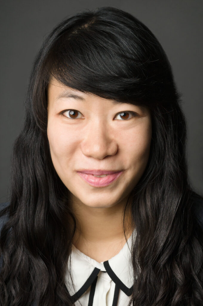 Headshot of a woman in her 20s who has heritage from South Korea and Laos she has light skin tone and long black wavy hair with swooped bangs. She is wearing a white button up with black edging. She is looking at the camera.