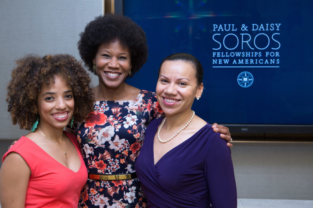 Photograph of three women standing in front of a screen that has the PDSoros logo on it. The woman in the middle, the eldest of the three is in her 50s has her arms around the other two, who are in their 30s. 