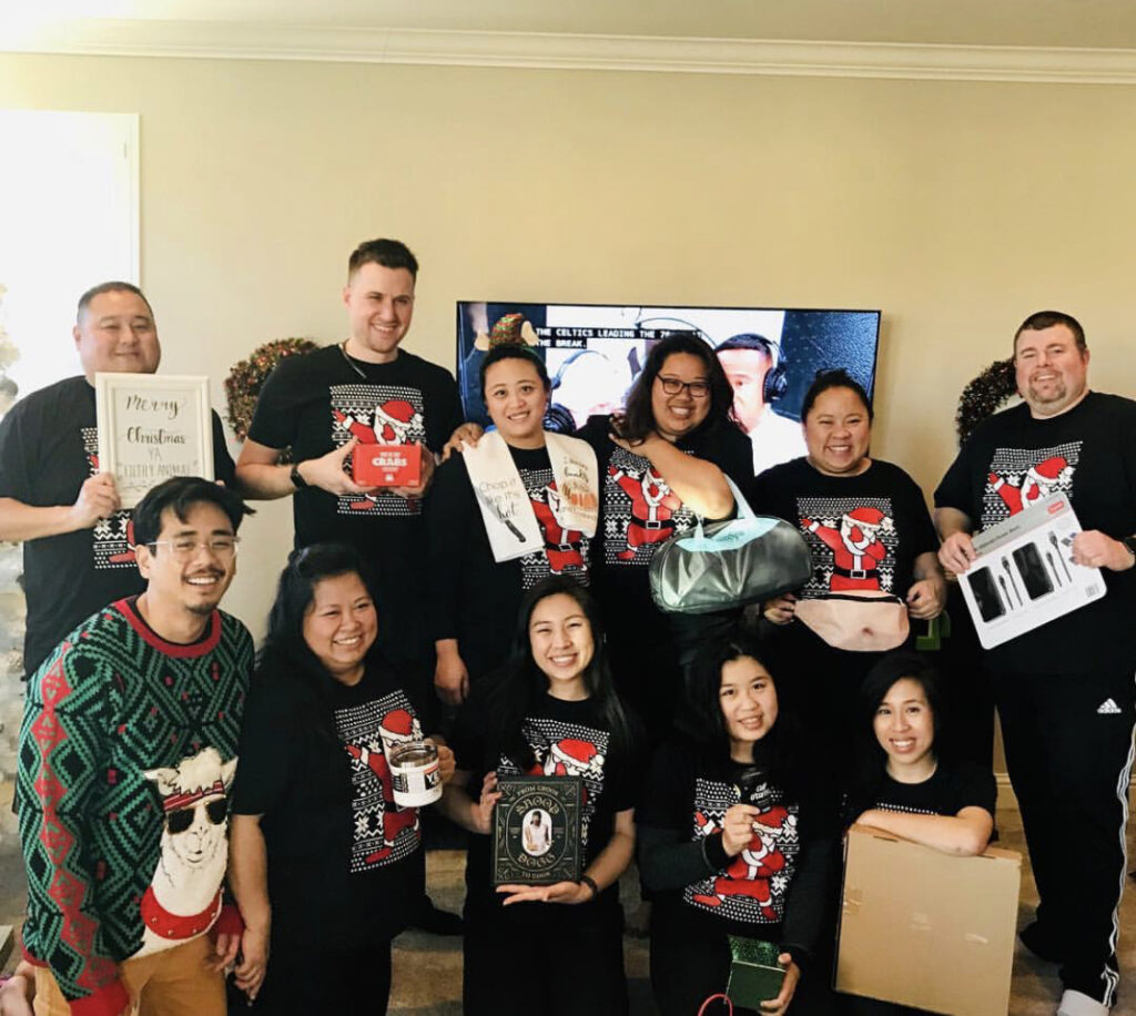 Photograph of a group of eleven people posing in two rows for the camera. Everyone is wearing a holiday themed sweater, most of them are wearing the same sweater. They are holding up what appear to be gifts. Everyone is smiling.  