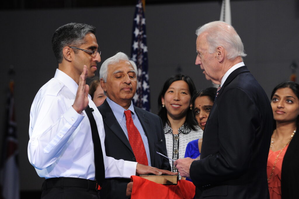 Photograph of a swearing in ceremony. The man being sworn in is in his 40s with medium skin tone and greying blacking hair, he is wearing a white button up shirt and black tie, his right hand is raised and his left hand is on a book. The man swearing in is in his 70s with light skin tone and white hair. 