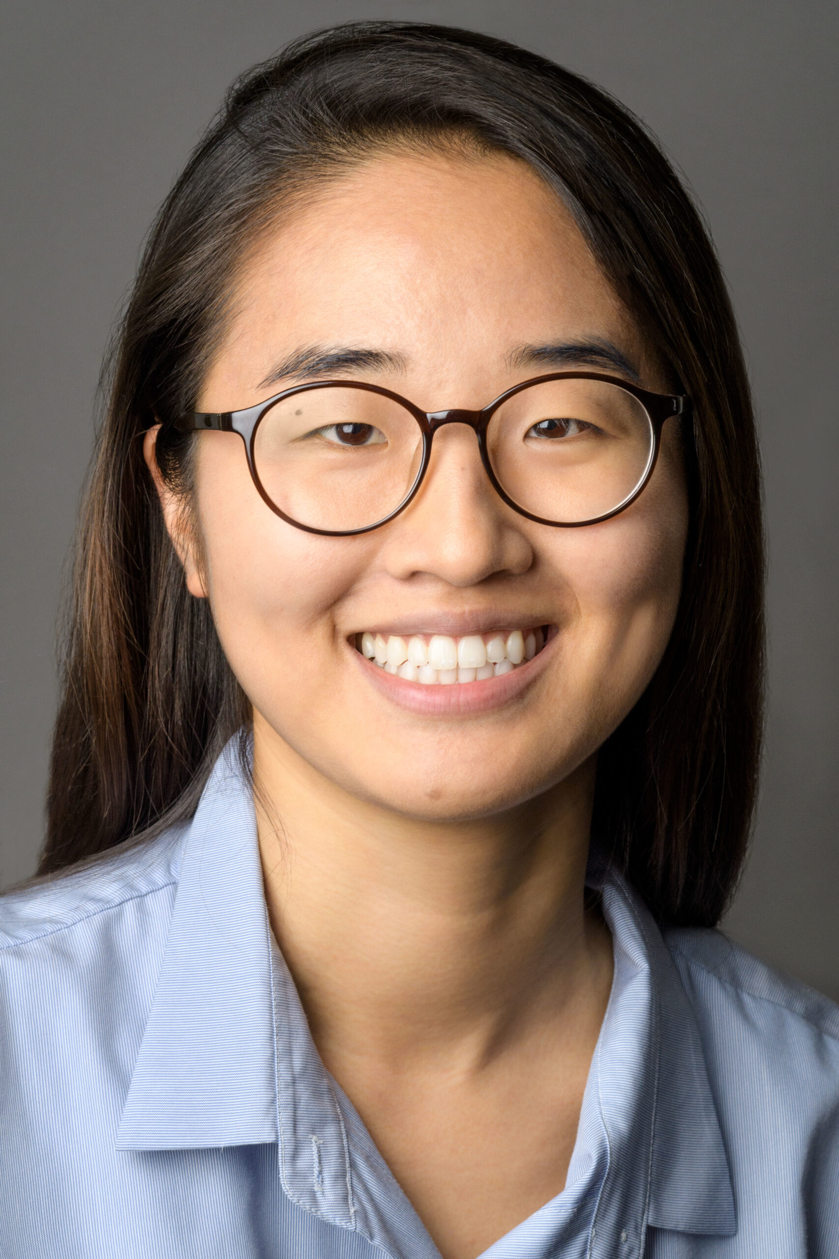 Headshot of a woman in her 20s who has heritage from South Korea, with medium-light skin tone and long straight black hair. She is wearing a light blue button up shirt and round black framed glasses. She is smiling at the camera. 