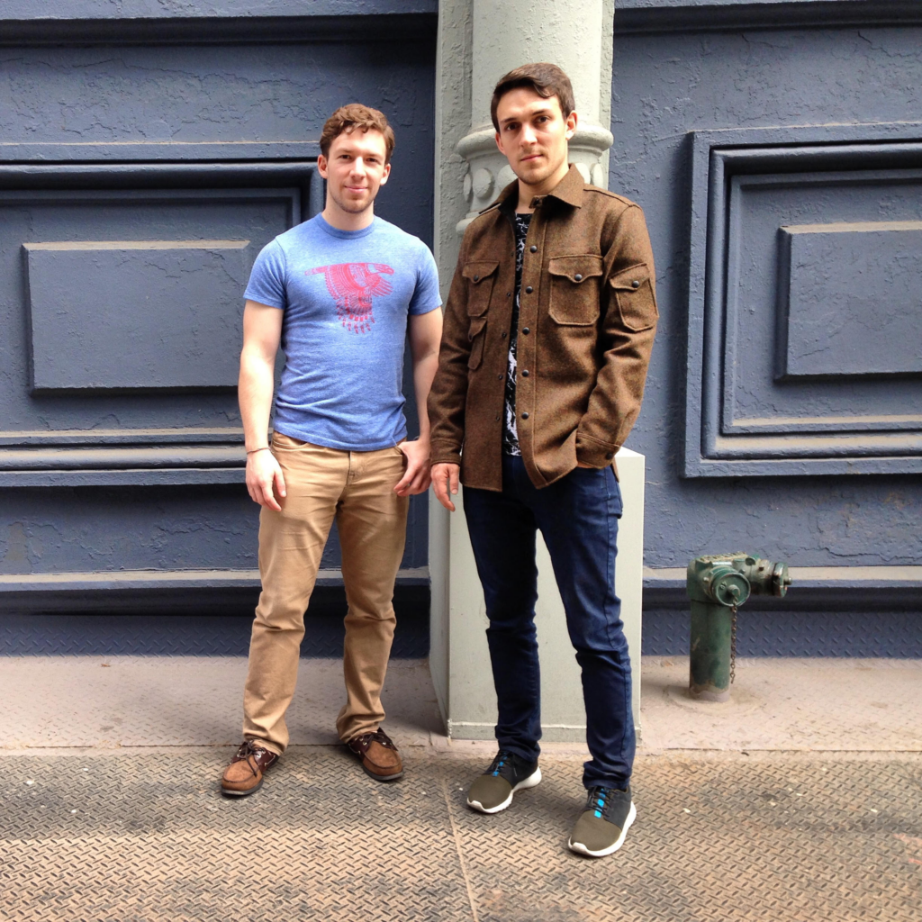 Photograph of two men in their 20s standing in front of a wall with built out pieces. 