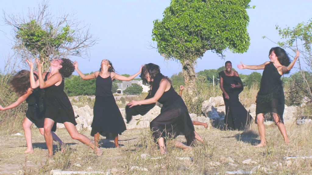 Photograph of people dressed in all black and appear in mid-dance, trees and other greenery can be seen around them. 