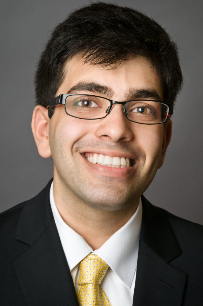 Headshot of a man in his 20s who has heritage from India with light skin tone and black hair. He is wearing a black suit, white button up shirt, yellow tie and black rimmed oval/rectangle glasses. He is smiling at the camera.