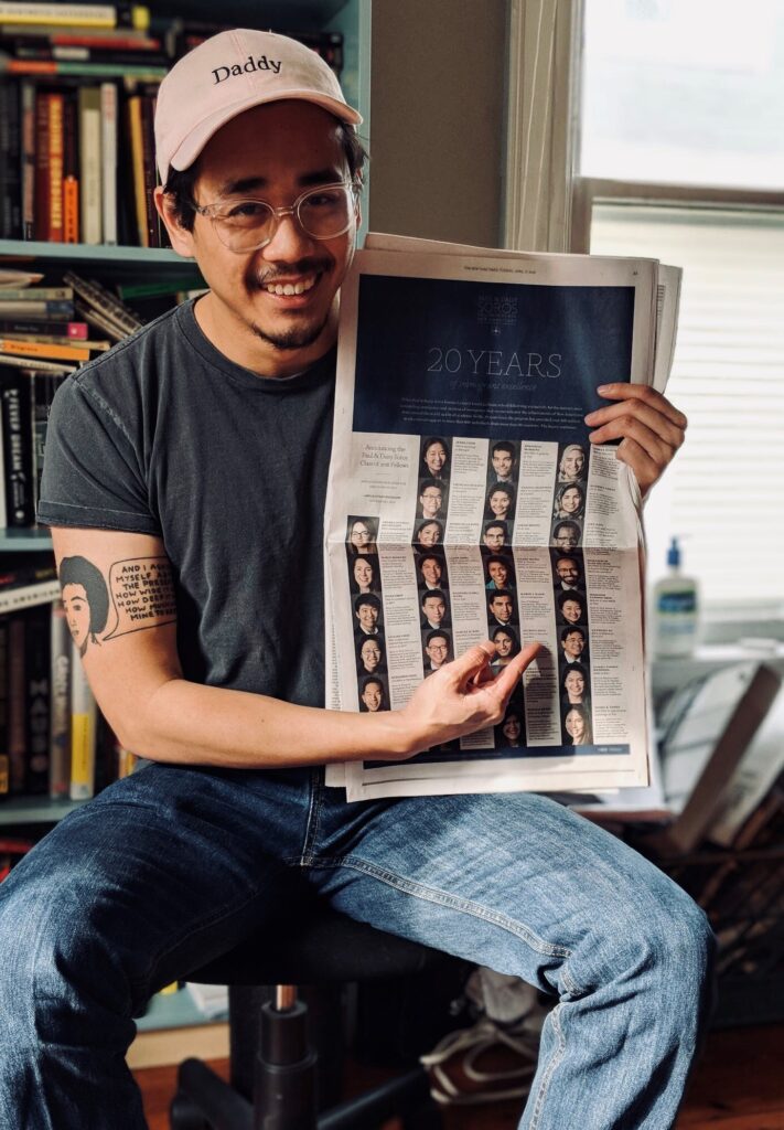 Photograph of a man in his 20s, he is wearing a grey t-shirt, jeans and a baseball cap that says "Daddy". He is sitting on a stool, a bookshelf and a window can be seen behind him. He is smiling at the camera. 