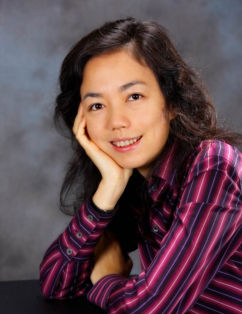 Headshot of a woman in her 20s who has heritage from China with light skin tone and dark brown curled hair. She is wearing a purple and pink striped button up shirt. She is seated and leaning forward with her arms crossed, one hand gently rests under her chin, she is looking at the camera and smiling.