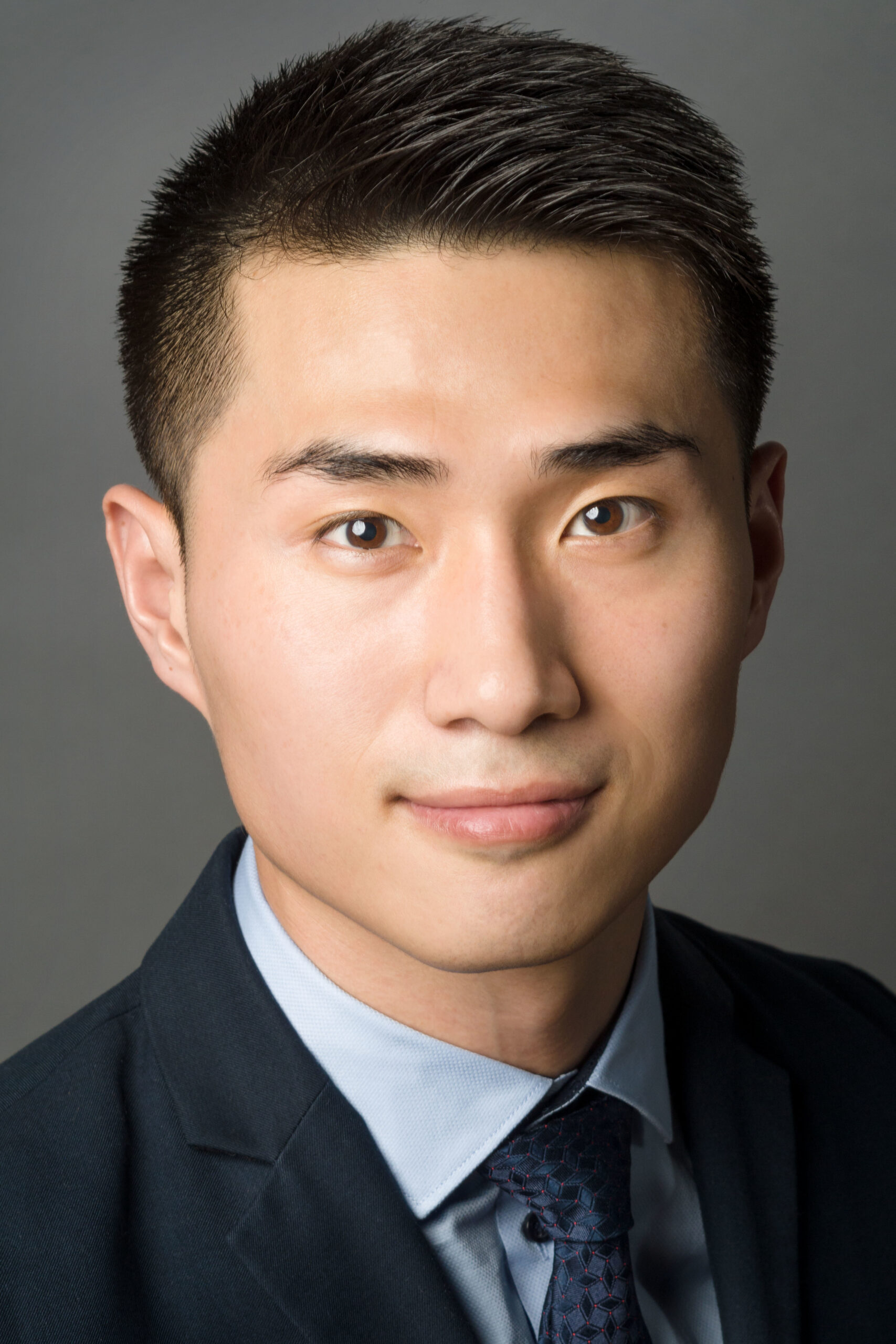 Headshot of a man in his 20s with light-medium skin tone and black short hair. He is wearing a navy suit, light blue button up and blue tie. He is looking at the camera.