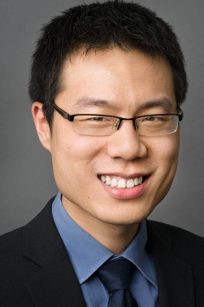 Headshot of a man in his 20s who has heritage from Taiwan with light-medium skin tone and black hair. He is wearing a black suit, blue button up shirt, navy tie, and half-rimmed narrow rectangle glasses. He is smiling at the camera.