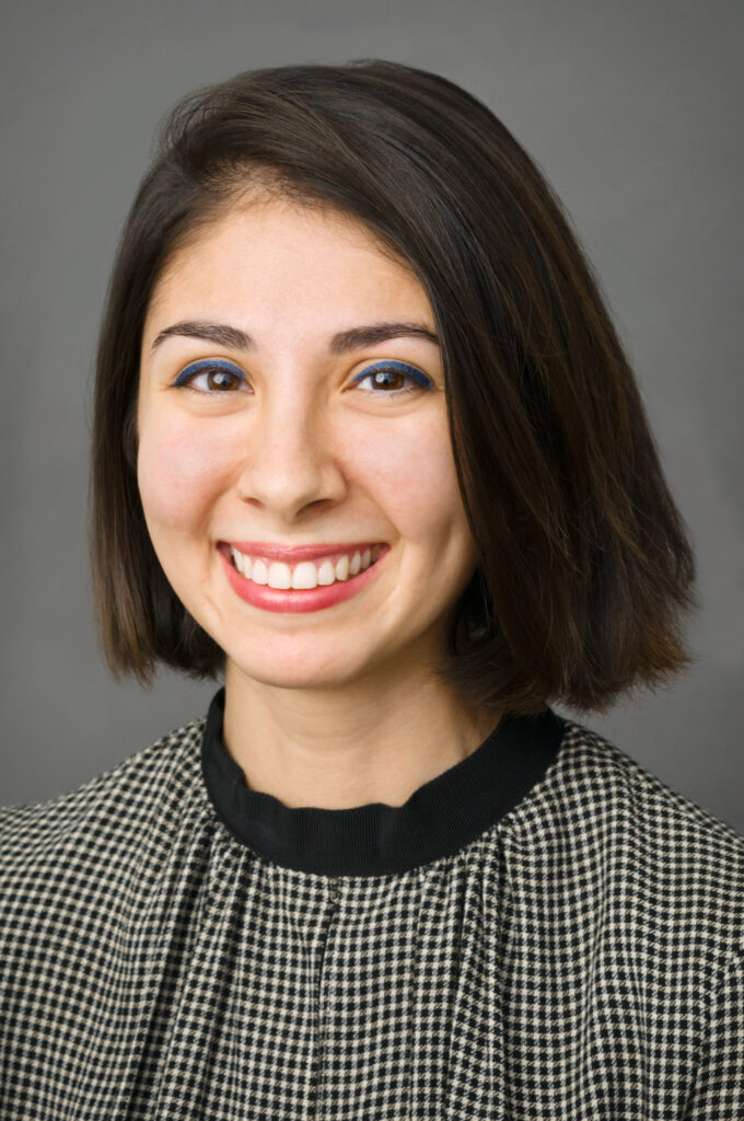 Headshot of a woman in her 20s who has heritage from United Arab Emirates, she has light skin tone and dark brown hair cut in a long bob. She is wearing a black and tan gingham patterned with a black collar. She is smiling at the camera.