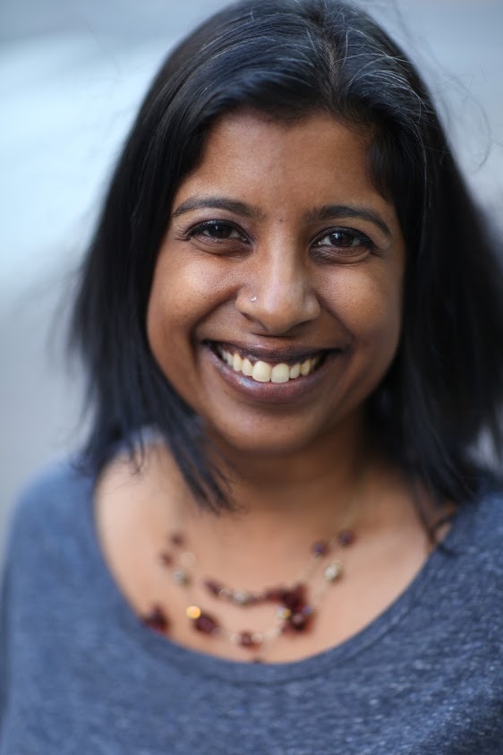 Headshot of a woman in her 40s who her heritage from India, she has medium skin tone and black shoulder length hair. She is wearing a blue top and red beaded necklace. She is smiling at the camera. 