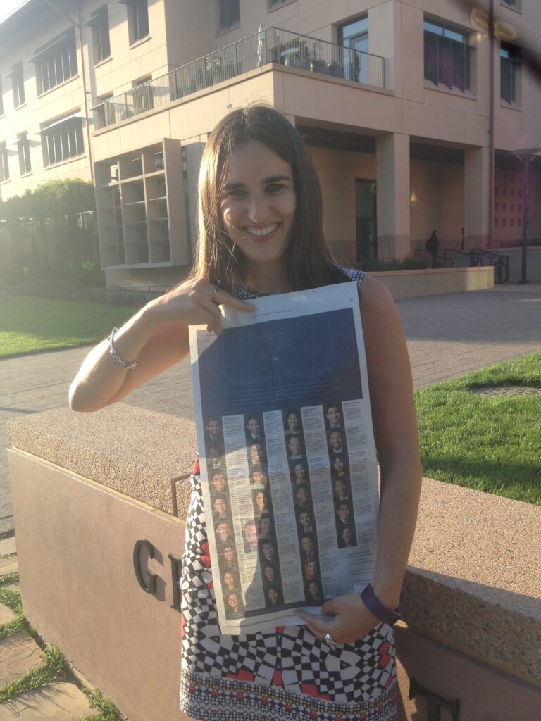 Photograph of a woman in her 20s who has  heritage from United Kingdom and Iran with light skin tone and long straight brown hair stands in front of a light colored building, sidewalk and lawns can be seen, holding a New York Times and smiling. 