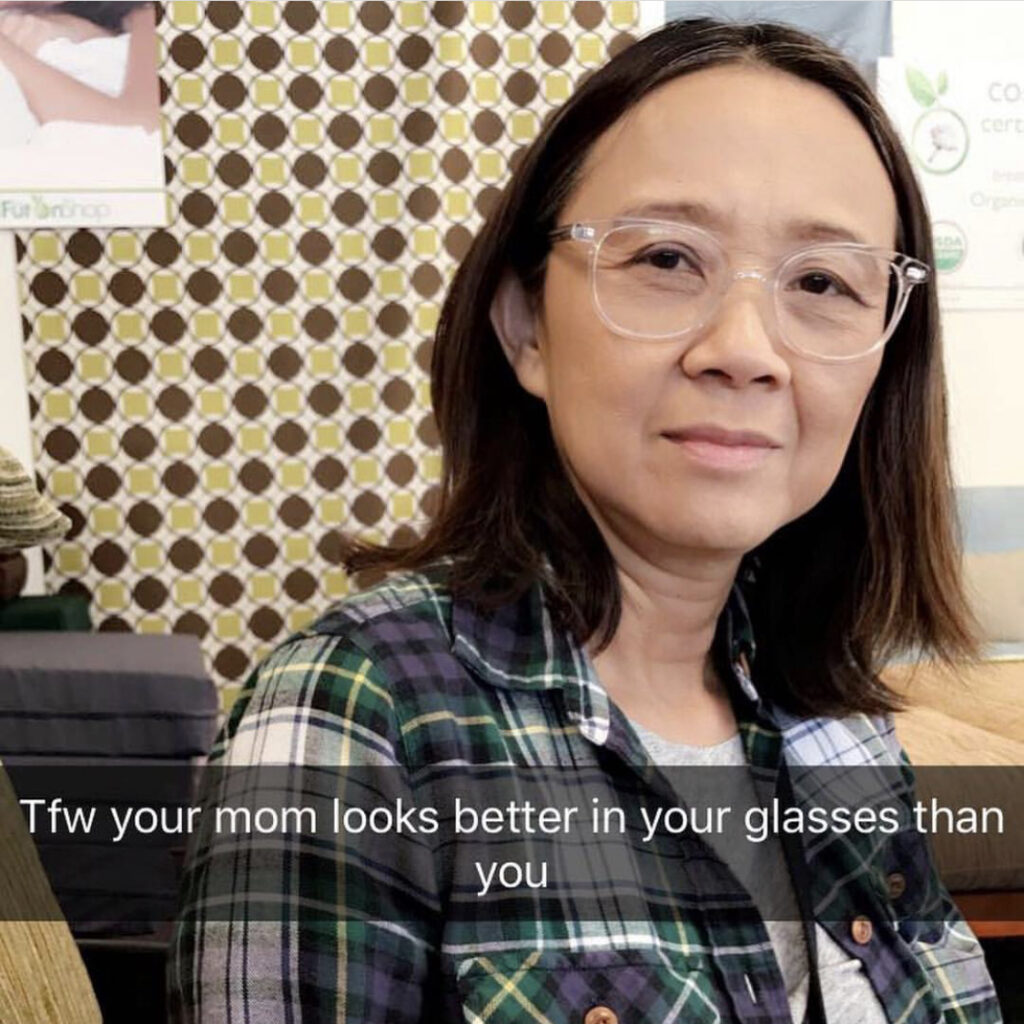 Photograph of a woman in her 40s with light skin tone and shoulder length black hair. She is wearing large clear rimmed glasses and a tartan pattern shirt. There is a caption that says "Tfw your mom looks better in your glasses than you".