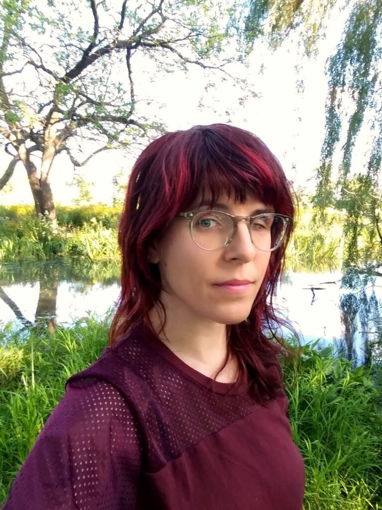 Photograph of a woman in her 20s who has heritage from Italy with light skin tone and dyed dark red / pink shoulder length hair with bangs. She is wearing a wine-red top and large clear rimmed glasses. Grass, trees and water can be seen behind her. 