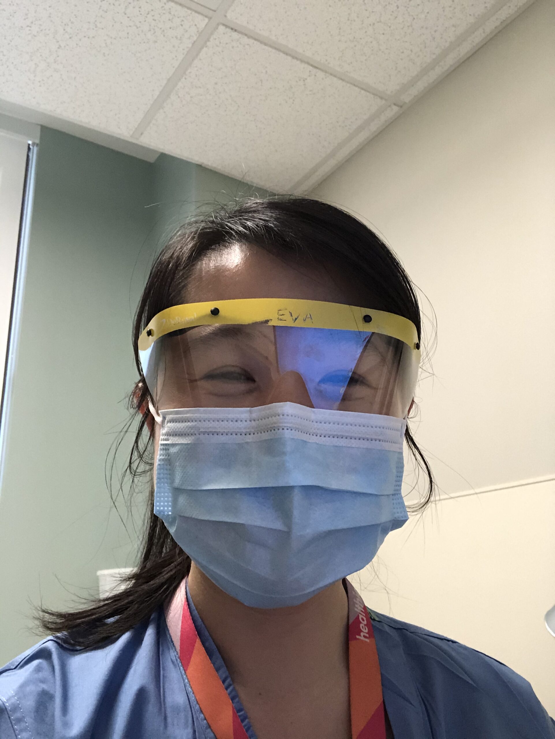 Selfie of a woman in her 20s who has heritage from China with light skin tone and long black straight hair pulled back with side bangs, across her left forehead. She is wearing a surgical mask and eye shield, blue scrubs and a lanyard.