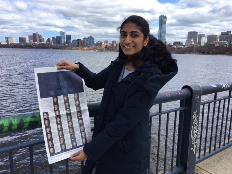 Photograph of a woman in her 20s who has heritage from India, with medium skin tone and pulled back black hair. She is standing at a railing overlooking a body of water, tall buildings can be see on the other side of the water. She is holding a New York Times and smiling. 