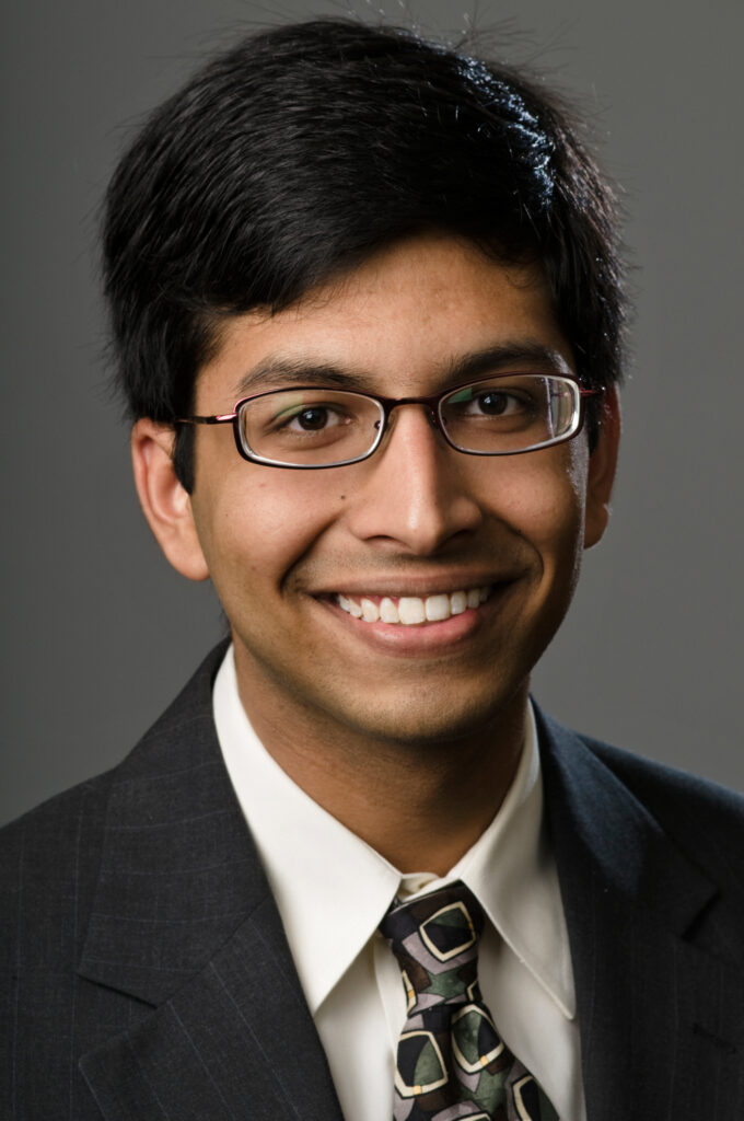 Headshot of a man in his 20s who has heritage from India with medium skin tone and black short hair. He is wearing a black suit, white button up shirt, multi-colored tie and black rimmed oval glasses. He is smiling at the camera.