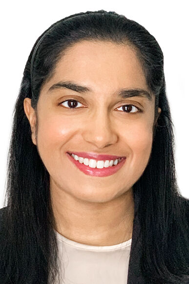 Close-up headshot of a woman in her 20s who has heritage from India with medium skin tone and long black hair half pulled back and a black headband; she is wearing a black blazer and a white blouse; she is smiling at the camera