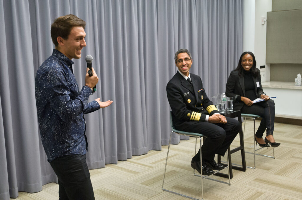 Photograph of three people, the man on the left is in his 30s with light skin tone and brown hair, he is wearing a blue button up shirt, black pants and is holding a microphone and gesturing to the other people in the picture. They are seated to his left in stools, they are both in their 40s. The man in dressed in a military uniform with medium skin tone and grey hair. The woman has medium-dark skin tone and black hair. 